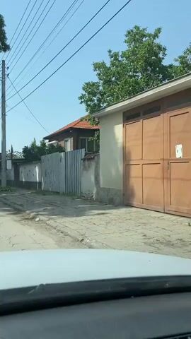 A car drives past a quiet street in a Bulgarian village on a sunny day. A Bulgarian flag is visible hanging from a house, and an old rug hangs on a clothesline in front of another house.