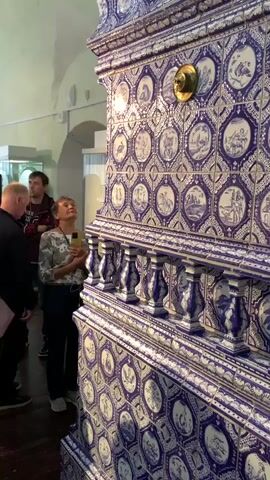 The video shows a group of people in a museum or gallery. The camera is focused on a large, ornate tiled stove or fireplace, predominantly blue and white, with intricate patterns and depicted scenes on its tiles. A woman in a patterned shirt and dark pants is standing in front of the stove, looking upwards and holding a smartphone, seemingly taking a picture or recording. Several other individuals, including men and women of various ages, are also present, some looking at the stove, others engaged with a tour guide who is speaking and gesturing towards a display case containing a portrait. The environment appears to be indoors, with wooden floors, arched ceilings, and bright lighting.