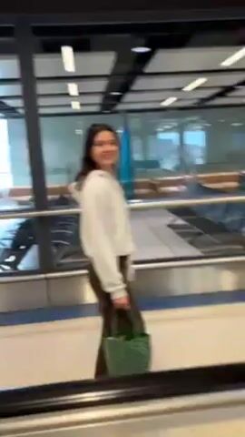 Here is a description of the video:
The video shows a woman in an airport terminal. She is wearing a white hoodie and brown pants. She is carrying a small green tote bag. She turns toward the camera and smiles, appearing excited. She walks on a moving walkway away from the camera and toward the terminal exit. The terminal is brightly lit with tile ceilings and glass walls. There are passengers in the distance.