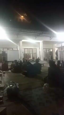 A group of people are sitting on the floor in rows, facing forward. They appear to be listening to someone speak, as there is a projector screen in the background. The setting appears to be an outdoor covered area, possibly a veranda or pavilion. The lighting is dim, with bright lights illuminating the area from above. It is nighttime, and the sky is dark. There are several tables in the foreground with cups and other items on them. The people are mostly dressed in dark clothing, and some are wearing hats.