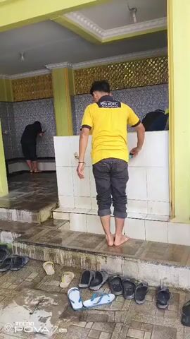 The video shows an outdoor area with a tiled floor and yellow columns. A man in a yellow t-shirt and dark pants is standing and facing a white tiled wall with faucets. He is washing his hands. In the background, two other individuals are also washing their hands at faucets. In the foreground, there are many pairs of sandals and flip-flops scattered on the wet floor. The environment appears to be a public washroom or ablution area, possibly associated with a mosque or religious institution.