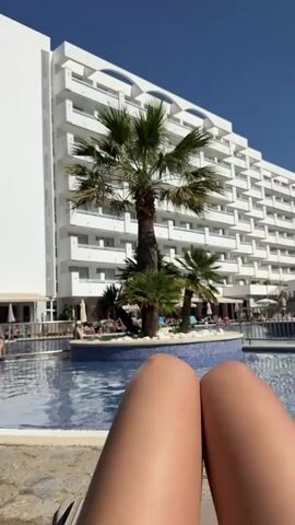 This media file depicts a sunny day at a resort in Es Camp de Mar, Spain. The scene is framed from the perspective of someone relaxing by a swimming pool, with their legs in the foreground. Beyond the pool, a large, modern white hotel building with multiple balconies dominates the background. Palm trees are scattered around the pool area, adding to the tropical ambiance.

Several people are visible around the pool, some lounging on sunbeds and others in the water. The water in the pool is a clear blue and reflects the bright sunlight. The sky is a vibrant, cloudless blue, indicating clear weather. The overall atmosphere appears relaxed and leisurely, suggesting a vacation or holiday.

The time of day is clearly daytime, likely late morning or early afternoon, due to the strong sunlight and long shadows. There is no visible text in the media file. The setting is unmistakably a resort with a swimming pool and a large hotel, characteristic of a vacation destination. The focus on the legs in the foreground, combined with the tranquil pool and resort setting, evokes a sense of relaxation and enjoyment.