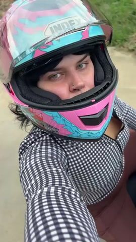 A woman wearing a pink, blue, and white patterned motorcycle helmet with a clear visor is sitting on a motorcycle. She has dark hair, a nose ring, and is wearing a black and white checkered long-sleeved shirt and brown pants. The visor of her helmet reflects trees and the sky. The video is filmed from a first-person perspective, looking down at the woman. The background appears to be outdoors with a paved surface and some greenery visible.