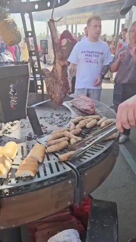 A man wearing a black baseball cap and a brown apron is grilling sausages on a large circular grill outdoors. He is using tongs to turn the sausages. To his left, there are slices of bread on the grill. Above him, a large tomahawk steak is hanging from a hook and cooking. In the background, other people are visible, and there are signs and awnings. The man in the apron appears to be interacting with someone to his right, who is gesturing. Another man in a white t-shirt with text on it is standing behind the grill. The environment appears to be a public event or food stall.