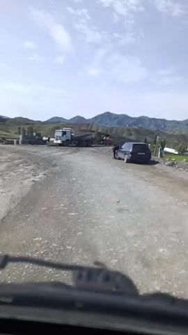 The video shows a dirt road with several construction vehicles, including a dump truck and an excavator. There are also some large concrete pipes stacked on the side of the road. In the background, there are mountains and a clear sky. There is one person standing near a car. The camera appears to be mounted inside a vehicle, looking out at the scene.
