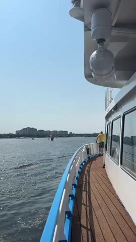 The video shows a person on the deck of a boat. The boat is moving on a body of water, and there are buildings and a bridge in the distance. The sky is clear and blue.