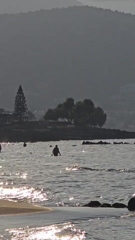 The video shows a beach scene with several people in the water. The sun is shining brightly, casting reflections on the water's surface. In the background, there are hills and trees. One person is wading in the water, facing away from the camera. Another person is swimming in the distance. Some rocks are visible in the water near the shore. The sand on the beach is visible in the foreground.