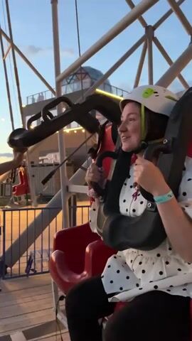 A woman wearing a white helmet with green accents and a black and white polka dot shirt is seated in a ride apparatus. She is holding onto the safety restraints with her hands and smiling. A person's arm is visible to her left, reaching out towards the apparatus. The background shows a metal structure, lights, and a dusky sky with hints of orange and blue. The scene appears to be an amusement park or similar attraction.