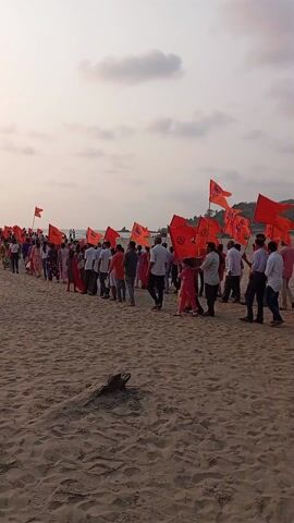 The video shows a crowd of people walking on a sandy beach. Most people are holding identical orange flags with a symbol on them, and are walking in the same direction. Some people are wearing white shirts and pants, while others wear colorful traditional Indian clothing. The sky is overcast and the ocean is visible in the background. There is a piece of driftwood in the foreground. The camera is mostly stationary.