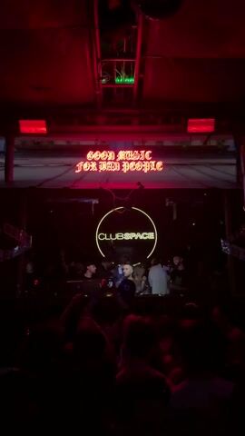 A vertical shot of a nightclub with a sign that says "GOOD MUSIC FOR BAD PEOPLE" in red neon. Below that is another sign, a glowing yellow circle that reads "CLUBSPACE" in the middle. In front of the signs are a crowd of people; most are blurred and in silhouette because of the dark setting and red lighting, but some figures are illuminated in front of the Clubspace sign. They appear to be dancing and enjoying the music. Above, some of the ceiling structure and other equipment can be seen.