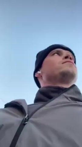 A man wearing a black beanie with white text on it, earbuds in his ears, and a grey jacket, looks up at the sky. Then he looks down at the camera and smiles. The background is a clear, light blue sky.