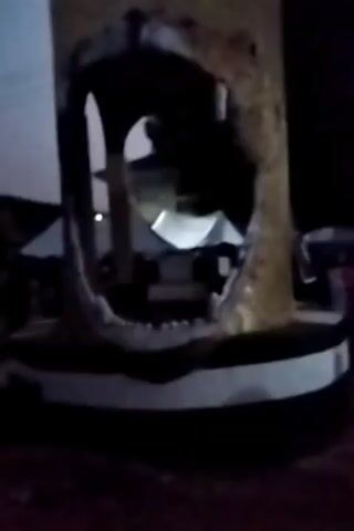 The video shows a cylindrical structure with the image of a lion's head and open mouth cut out from it. The structure is set on a circular base painted with a black and white stripe. Behind the structure, in the open mouth area, is a building with a white roof. The sky is dark, suggesting that the video was taken at night. There are also hints of other structures and people in the background, but they are largely obscured by the darkness.