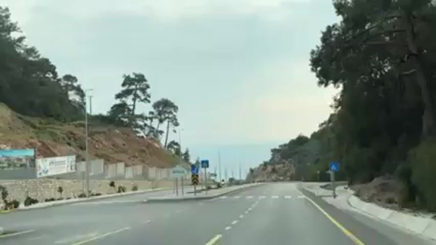 A car travels down a four-lane road with mountains on either side. A pedestrian crosswalk can be seen. The road is bordered by sidewalks and a gray stone wall. A motorcyclist passes by on the left. The ocean can be seen in the distance. The sky is cloudy.