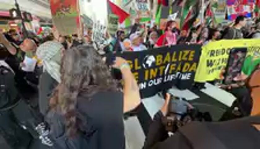 This video depicts a large outdoor protest or demonstration taking place on what appears to be a city street during the daytime. A dense crowd of people, diverse in age and attire, are seen marching forward. Many participants are holding Palestinian flags, featuring red, black, white, and green colors, which are frequently visible throughout the footage.

The atmosphere is energetic, with individuals actively moving and chanting. The audio captures a repetitive chant, strongly suggesting a call for "Intifada." Several banners and signs are prominently displayed. A large yellow banner at the beginning of the video clearly reads "GLOBALIZE THE INTIFADA," accompanied by a graphic of a raised fist. Another sign, partially visible, contains the phrase "F*CK NYPD." A different sign, briefly shown in the middle of the video, states "LET'S MAKE A NEW WORLD."

The lighting indicates it is daytime, and the general appearance of the crowd suggests a determined and unified demonstration. The background shows blurred urban structures, consistent with a street setting in a city like New York, United States.
