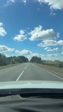 A vertical, first-person perspective video shows a view from inside a car driving on a paved road. The sky is a vibrant blue with scattered white, fluffy clouds. The road ahead stretches into the distance, bordered by green trees and fields. White lines mark the lanes on the road. The dashboard of the car is visible at the bottom of the frame. The sun appears to be shining.