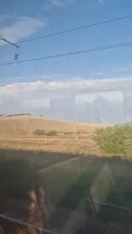 The video shows a landscape viewed from a moving vehicle, likely a train, as indicated by the train tracks visible at the bottom of the frame. The scene is dominated by a clear blue sky with scattered white clouds. The terrain is predominantly flat with rolling hills in the distance. The landscape is a mix of dry, golden fields and patches of green vegetation, including trees and bushes.

Several small structures, appearing to be houses or outbuildings, are scattered across the landscape. Some have white walls and darker roofs, while others appear to be in a state of disrepair. Utility poles and wires are also visible, suggesting the presence of infrastructure.

Reflections of the interior of the vehicle are visible on the windows, partially obscuring the view. In the foreground, a blurred reflection of a person's face and shoulder is visible on the left side of the frame. The lighting suggests it is daytime, likely late afternoon or early morning, given the quality of light and the shadows. The overall impression is of a rural, perhaps sparsely populated, environment.