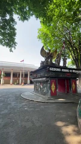 The video shows a wide-angle shot of a paved outdoor area in front of a building. On the right side of the frame, there is a dark, multi-tiered monument with statues and ornate carvings. The monument has the words "GEDUNG JUANG 45 NGANJUK" written on it in red and gold letters. Red and gold decorative elements are visible on the sides of the monument, as well as what appears to be a relief carving of a scene with a flag. To the left of the monument, a long, white building with a red roofline and columns is visible. The building has the same text, "GEDUNG JUANG 45 NGANJUK", written on it. A flagpole with an Indonesian flag is situated in front of the building. Lush green trees with dense foliage frame the top and sides of the shot, casting shadows. The ground is an asphalt surface with some cracks and wear. In the background, two individuals on motorcycles are present, one on the left and one further back in front of the building. The sky is clear and bright, suggesting daytime.