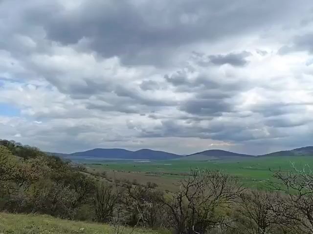 The video shows a landscape with a cloudy sky. In the foreground are trees with bare branches. Beyond the trees, a green valley stretches out. In the distance, hills or small mountains can be seen against the horizon. The sky is filled with gray clouds.