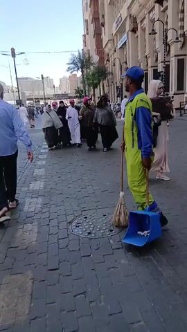 A man in a bright yellow and blue uniform with a blue cap sweeps a paved street with a broom and blue dustpan. He glances at the camera as he pauses near a manhole cover. A small crowd of people wearing traditional clothing is visible walking behind him. Other people in casual clothes walk in the opposite direction. Buildings are seen on either side of the wide street, and street lamps are overhead.