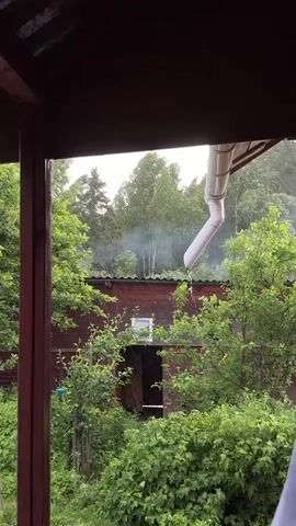 The video is a POV shot from inside a porch, looking out at a wooded area. In the foreground, there are dark red wooden beams supporting the roof of the porch. Green trees and bushes fill the sides of the frame. In the middle ground, there is a dark red wooden building with a white downspout extending from the roof. A small, open doorway is visible on the building, with dark fabric hanging inside. There is a wisp of white smoke rising from behind the building, near the roofline. In the background, there are more trees, some with light-colored bark, and a cloudy sky. The downspout is collecting water and appears to be raining or drizzling.