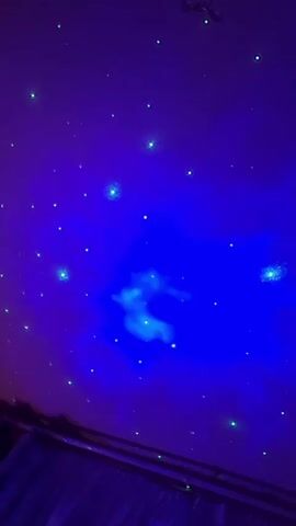 A starry night sky projection is displayed on a ceiling. The projection features a blue nebula-like cloud formation in the center, surrounded by numerous green and white stars. The overall lighting is dark, with the blue and green lights of the projection being the primary light sources. The bottom of the frame shows a dark, possibly curtained area, suggesting an indoor setting. The video appears to be a static shot of this projection.
