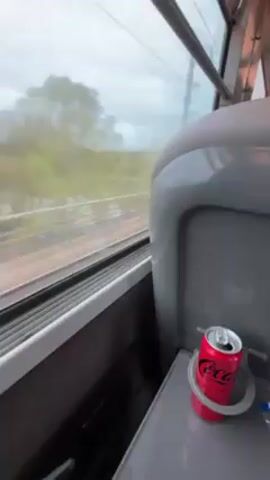 The video shows the view from inside a moving train. Outside the window, the landscape passes by, featuring fields, trees, and an overcast sky. A can of Coca-Cola sits in a cupholder attached to the train seat. A hand is visible near the bottom of the frame, resting on the surface next to the cup holder.
