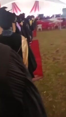 The video appears to show a graduation ceremony taking place inside a large tent. The camera focuses on a line of graduates dressed in black gowns and mortarboards, some with colorful stoles. They are standing on a red carpet placed on a grassy surface. In the background, there are rows of chairs and tables covered with red tablecloths, presumably for guests. A screen is visible at the far end of the tent, suggesting presentations or speeches are part of the ceremony. The overall atmosphere seems to be celebratory and formal.