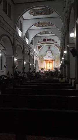 The interior of a church is depicted, filled with rows of dark wooden pews populated with people. Arched ceilings extend down the length of the building, with paintings visible in the arches above the altar. The altar area is well-lit, featuring a cross and banners labeled "SAN CALIXTO" and another word, perhaps an alternate banner for San Calixto. People are mostly seated and some are standing in the background. There are spherical lamps on the pillars beside the aisles and some speakers mounted on the pillars. The camera is positioned in the back of the church, looking toward the altar.