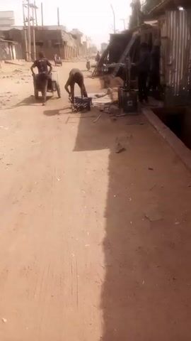 The video shows a street scene, seemingly in a somewhat impoverished area. The ground is dirt, possibly a road, with visible tire tracks. A person bends over a blue toolbox on the ground, working on metal pieces, possibly constructing something. Nearby is a welding machine and a pile of various metal pieces. Further down the road, another person is pushing a cart with another figure standing nearby. The area has basic structures and buildings on both sides of the road. The overall impression is one of manual labor being performed in a somewhat informal setting.