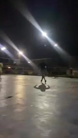 A soccer game is being played on a paved court at night, illuminated by overhead lights. Several people are visible, mostly in silhouette, running around and kicking a soccer ball. The court is surrounded by a metal fence, and there are some trees visible in the background. The camera is handheld and moves slightly as it records the scene. The overall impression is one of an informal, after-dark game of soccer.