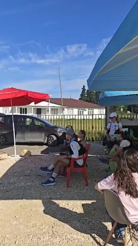 The video shows an outdoor event with several people standing around and a blue inflatable archway in the distance. In the first clip, two red pop-up tents are visible, with several people standing under them. There's also a dark-colored car parked nearby. In the second clip, the camera pans to reveal a dirt path leading to the blue archway, which has "ФИНИШ" written on it. Red and white flags line the path. Several people are walking towards or standing near the archway. The sky is clear and blue.