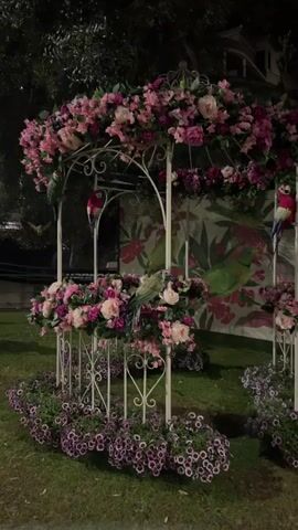 The video shows a white, ornate gazebo on a grassy lawn at night. The gazebo is adorned with various pink and purple flowers. Four colorful, artificial parrots are placed on the gazebo. Behind the gazebo is a wall with a floral and bird design painted on it. Small purple flowers are planted around the base of the gazebo. The scene is somewhat dark, illuminated by artificial light. A tree is visible in the background.