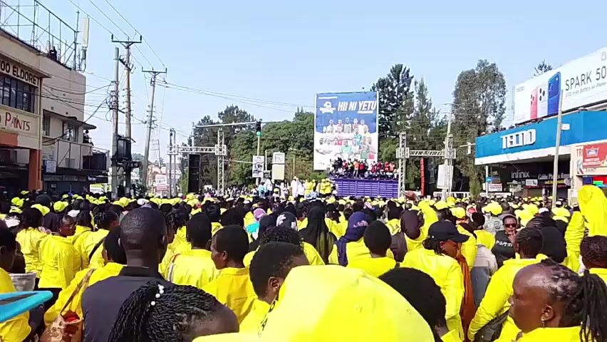 A large crowd of people, most of whom are wearing yellow shirts or jackets, are gathered in an outdoor urban area. They are facing a raised stage with a large banner that has text and images on it. Some people in the crowd are raising their hands. There are buildings, streetlights, and utility poles visible in the background. The sky is clear and bright, suggesting it is daytime. The overall scene suggests a public event or rally.