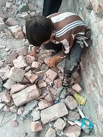 A boy in a striped shirt and dark pants squats among broken bricks, apparently searching for something. He is wearing sandals. A man stands nearby, also in sandals and dark pants. They are standing on a rooftop or next to a low brick wall with rubble. There is trash amongst the bricks. The lighting is bright.