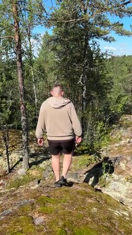 A person is walking down a rocky, moss-covered path in a forest. The person is wearing a beige hoodie and black shorts. The path is surrounded by trees and rocks. The sun is shining and the sky is blue.