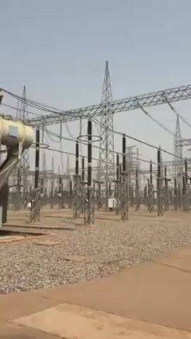 The video shows an electrical substation.  The substation is filled with transformers and towers connected by overhead power lines. The ground around the equipment is covered with gravel and there are concrete walkways. The sky is light blue. A motorcycle is parked near a small building.
