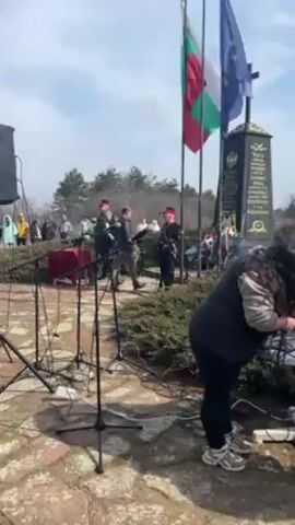 A low-angle outdoor shot shows a monument with the flags of Bulgaria and the European Union behind it. There is a small crowd of people in the background. In the center foreground, a woman with long dark hair, wearing a black vest over a white shirt, and black pants, is bent over a table, adjusting wires of sound equipment. There are several microphone stands in the left foreground. Some of the people in the background are wearing ceremonial garb. The scene takes place on a stone-paved area with some greenery. The sky is a light blue with some clouds.