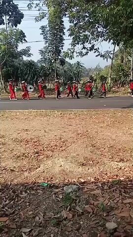 A marching band consisting of at least ten individuals in red uniforms with white hats and sashes is walking on a paved road, some carrying drums. One person in a white shirt and helmet is riding a motorcycle ahead of the band, seemingly escorting them. The scene is outdoors with trees lining the road and a dirt and leaf-covered area in the foreground. The sky is visible and appears to be daytime.