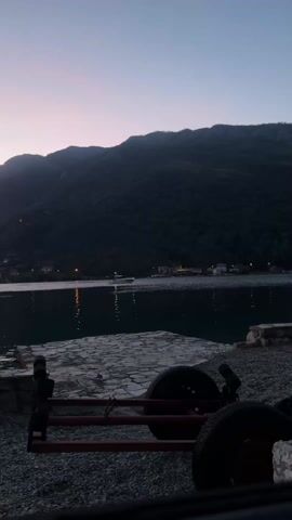 The video shows a lake surrounded by mountains during twilight. The sky is a blend of pink, orange, and dark blue. Lights are visible along the shore of the lake, and their reflections shimmer on the water's surface. In the foreground, there is a gray stone platform and a black and red object with wheels. The entire scene is somewhat dimly lit, emphasizing the darkening sky.