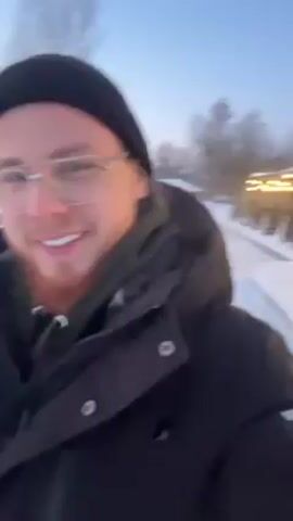 A man with a red beard, wearing glasses and a black beanie, smiles into the camera as he walks on a snowy path. He is wearing a black winter jacket. There are trees in the background and lights from a building on the right. The sky is a light blue.