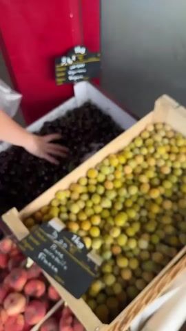 The video shows a close-up, slightly blurry view of a fruit stand. In the foreground, there are two wooden crates filled with fruit. The crate on the right is filled with small, round, yellow-green plums. The crate to the left of that is filled with dark red cherries, and a hand is reaching into the cherries from the left side of the frame. Below these two crates, a third crate contains red and pink peaches. Each crate has a black sign in front of it with white writing, indicating the price and type of fruit. Behind the fruit stand, a red surface is visible, possibly a refrigerator or wall, with another black sign attached. The lighting suggests an outdoor or well-lit indoor market environment.