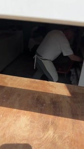 The video shows an older man handing a piece of bread through a window. The man is bald and wears a white t-shirt and white apron. He holds the bread, which is golden brown, in both hands and places it on a wooden surface in front of the window. The environment appears to be a bakery or food stall, with other food preparation items visible in the background. The lighting suggests it's daytime.