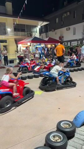 The video shows a group of children riding bumper cars in an outdoor amusement area at night. The bumper cars are brightly colored, with red, blue, and black vehicles visible. The children are actively steering and bumping into each other as they navigate the track, which is lined with tires. Spectators, including adults and other children, are standing around the perimeter of the track, watching the activity. In the background, there are buildings illuminated with lights, suggesting a commercial or entertainment district. The overall atmosphere appears festive and lively, with a sense of fun and engagement among the participants.