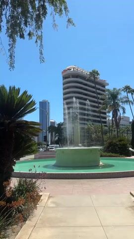 A vertical video captures a view of a fountain in a park or public space. The fountain, with a green basin, is in the foreground, with water spraying upwards. Behind the fountain, there is a light-colored, curved building with multiple floors and balconies, possibly an office building or hotel. To the left of this building, a taller, slender blue building is visible. Palm trees and other lush green foliage surround the fountain and the buildings, suggesting a warm climate. The sky is clear and blue. In the background, traffic can be seen on a road, indicating an urban setting. The video is short, lasting only a few seconds, and the camera appears to be stationary.