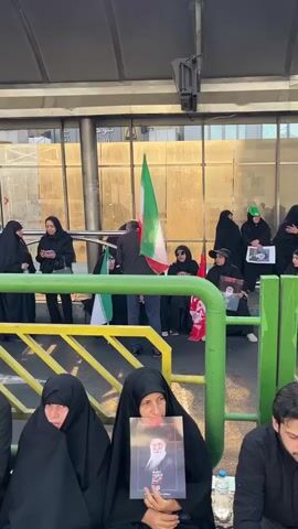 The video shows a group of people, mostly women wearing hijabs, gathered outdoors, sitting and standing behind a green and yellow barrier. An Iranian flag is visible, held by someone in the crowd. Some individuals are holding photographs. In the background, there is a building with glass windows and an ambulance with the word "AMBULANCE" visible. The ground appears to be paved. Some people are looking in the same direction, suggesting they are observing an event or procession. The atmosphere appears somber.