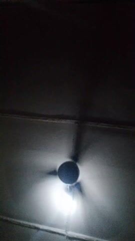 The video shows a dimly lit room with a ceiling fan spinning.  The light fixture attached to the fan is illuminated, casting a bright light downward. The camera angle is from below, looking up at the fan.  The ceiling appears to be painted or textured. The edges of the ceiling are visible, with what appears to be a molding or seam running along them. The rest of the room is dark.