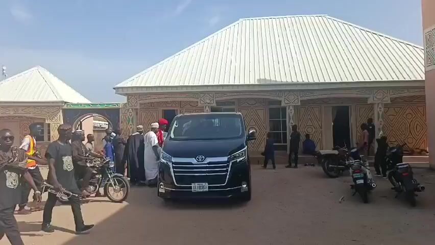 The video shows a gathering of people around a dark-colored Toyota van parked in front of a building with a white roof and decorative patterns on its walls. Several men are dressed in traditional attire, including a man with a red hat. Some people are standing near motorcycles. One man is carrying a trumpet. The setting appears to be a dusty courtyard with another similar building in the background. The sky is clear and blue.
