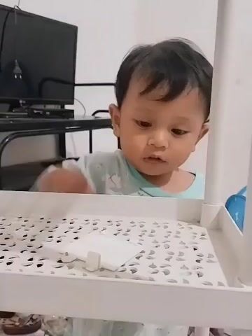 A young child with dark hair is sitting and looking at a white shelf. The child is wearing a light blue and white shirt with a pattern. The shelf has a white perforated surface. The child's right hand is on the shelf, and the child appears to be touching the surface. The child's mouth is slightly open. In the background, a dark television screen is visible on a stand, and a white wall is behind the child. The lighting is neutral, suggesting an indoor environment.