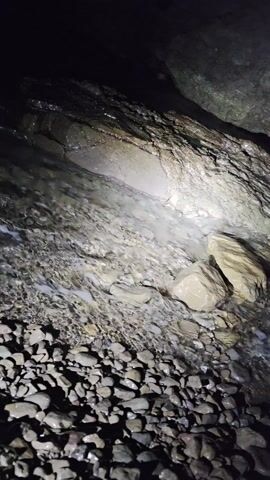 The video shows water flowing in a dark, rocky stream. The stream is shallow and contains small, dark rocks, with a few larger, lighter-colored rocks. The water is clear and flows quickly. The scene is dark, but a light source illuminates the water and the rocks directly beside it. There are also rocky walls on either side of the stream, further indicating the stream's location in a cave or underground setting.