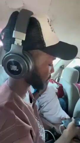 The video shows a person seated in a car, wearing a cap and JBL headphones. They are looking at a smartphone in their hands. Other passengers are visible in the background. The car appears to be in motion.